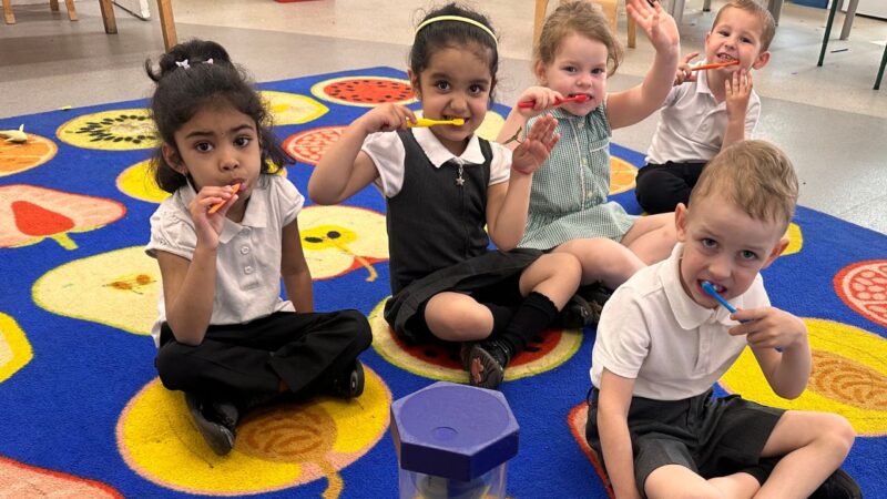 Five early years pupils at KIngs Oak Primary brushing teeth seated on mat as part of supervised toothbrushing programme