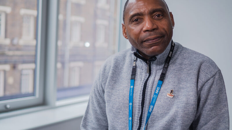 Headshot of Kirt Hunte, a Mental Health Nurse and Psychotherapist. He is looking straight at the camera and smiling. He is wearing a grey fleece and an NHS lanyard.